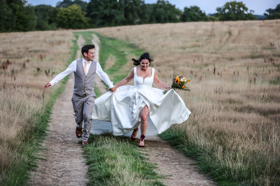 Bournemouth wedding photographer capturing bride and groom running hand in hand through countryside at sunset